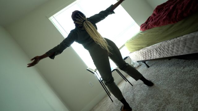 a dark skinned woman with long hair thrusts her chest toward the ceiling of a nearly empty bedroom