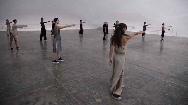 twelve dancers stand on an elevated platform with their right arms extended and index fingers pointing to a location out of frame