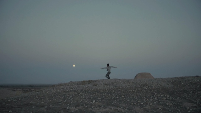 from a distance, at dusk, we see a dancer facing the moon, arms extended out to the sides and knees bent in parallel