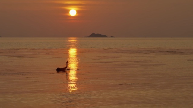 a lone person floats in the calm ocean at sunrise, their forearm extended toward the sky