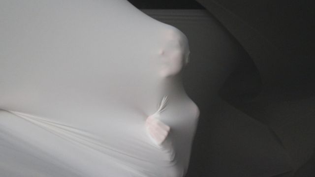 a person's face and left hand press into a wall of thin white fabric, lit harshly to accentuate the shadows