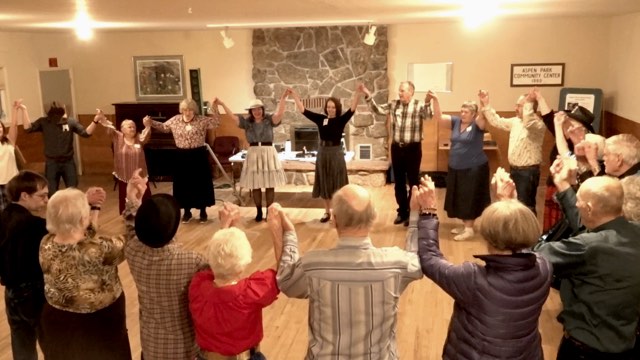a group of twenty older adults stands in a circle in large room in front of a brick fireplace, holding hands