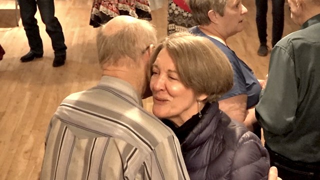 an older man and woman dance closely together in a room full of others; we see the woman's face, smiling
