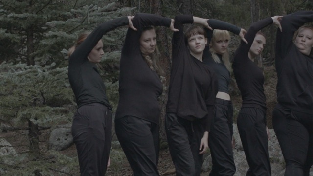 Six light skinned women of varying ages, all wearing black, are lined up with their right arms reaching over their heads, touching the right arm of the dancer to their left