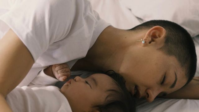 a woman with buzzed hair lays on a bed holding an infant; both have their eyes closed and are wearing white