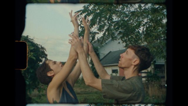 an image on a film strip of two light skinned dancers with short brown hair in a field in front of a house; one is helping the other raise their arms above their head