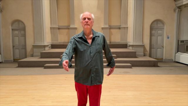 an older man in bright red slacks on a wood dance floor with a tiered stage behind him extends his right arm slightly in front of him and his left arm slightly behind him