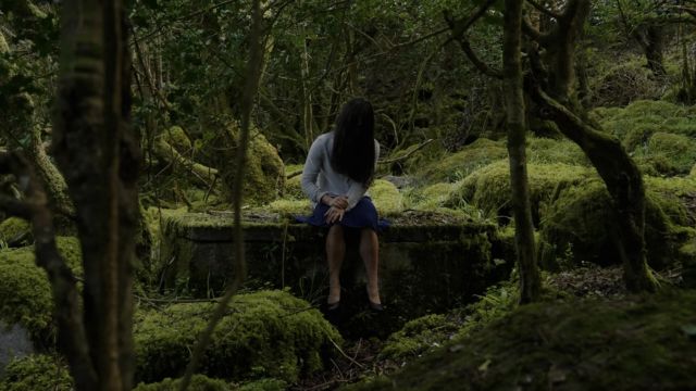 a woman sits on a flat concrete structure in the middle of a moss-covered forest, her long dark brown hair completely obscuring her face