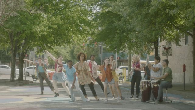 seven dancers and three musicians, all wearing bright colors, joyfully dance and play in the street of an urban area in bright daylight