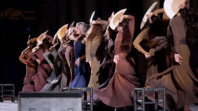 centered between eight spinning flamenco dancers wearing brown flamenco dresses and holding pericóns, an older woman in a blue dress stands singularly