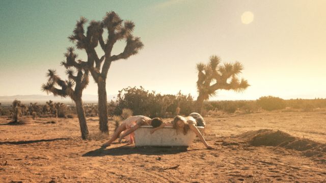 two women lie draped over a dirty bathtub in the middle of a desert environment; the sun and two Joshua trees are clearly visible behind them