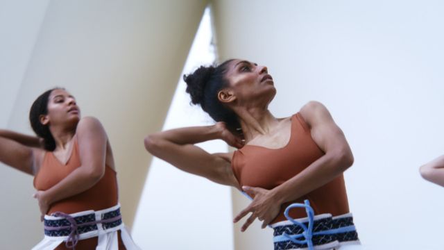 two dark haired, caramel skinned dancers in rust colored tank tops clutch their right ribs with their left hands