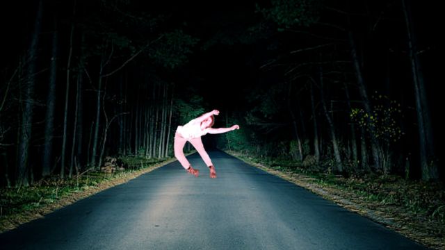 a grainy image of a dancer is superimposed onto a narrow dark road through the woods that is only illuminated by the headlights of a car outside the shot