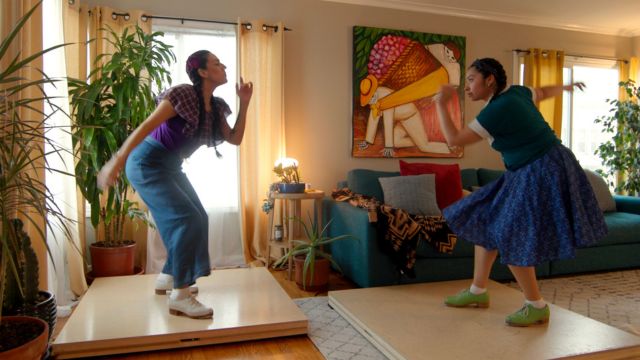 in a living room filled with large plants and bold colored art and furniture, two women on tap boards tap dance, facing each other