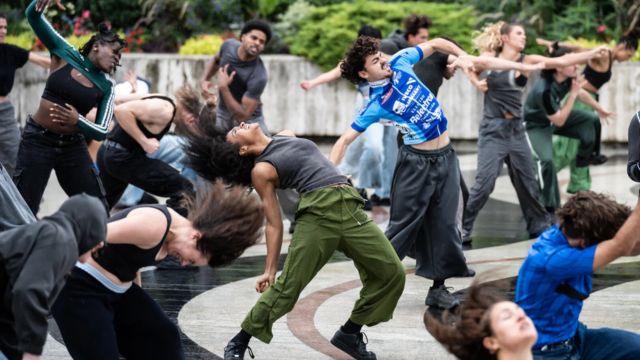 over a dozen younger dancers of various ethnicities dance and contort themselves in a public park