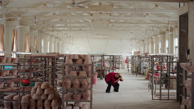 in a large white warehouse filled with shelves of pottery, a woman in a red shirt squats down intensely, holding her head