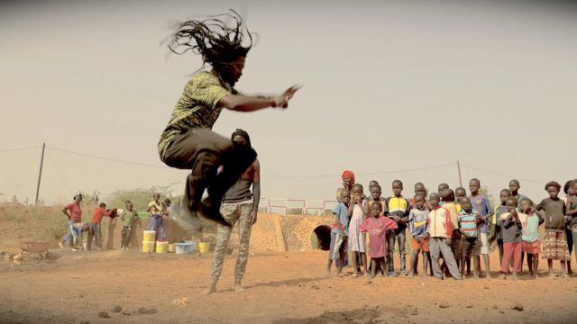 a dark skinned man with long dreadlocks leaps high in the air, knees tucking toward his chest, as over a dozen children gathered in a crowd look on