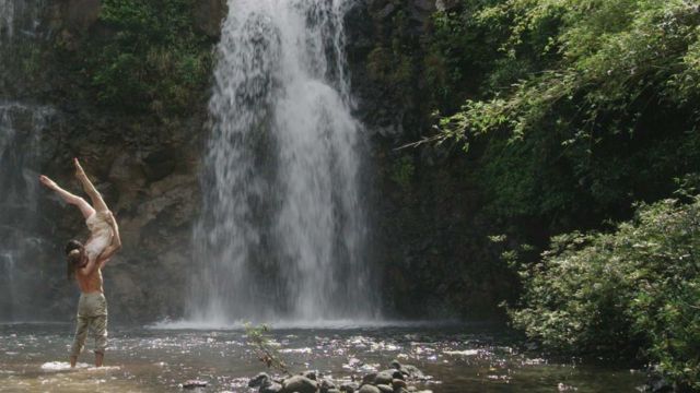 in a shallow pool at the bottom of an impressive waterfall, a dancer holds a female dancer upside down atop his right shoulder, her legs high in the air above her