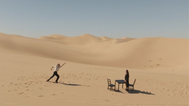 in a vast sea of sand dunes, a man strides exaggeratedly toward a dancer in all black, standing at a simple square table with two chairs
