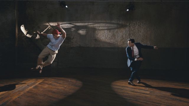two versions of a light skinned man - one wearing a tee shirt and baseball hat and one wearing a full suit and tie - dance in an empty room with a hardwood floor