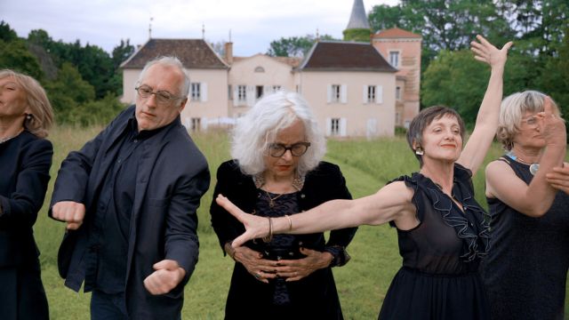 five older adults in black formal wear dance disparately in the foreground as a light colored two story manor appears in the background
