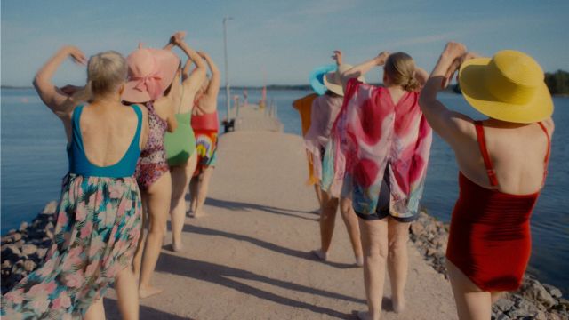 eight older adults wearing swimwear walk along a rock jetty, away from the camera and toward a large body of water, arms waving above them