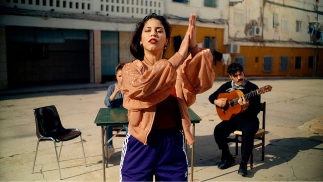 on a street in bright daylight, a young woman with dark hair and bright red lipstick doing flamenco dance; behind her and slightly out of focus are two people, one with a guitar, sitting at a table