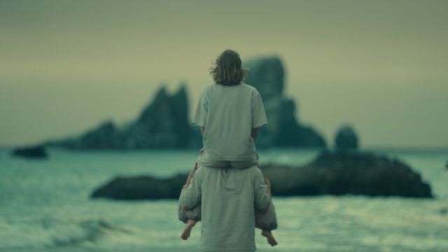 a dancer sits on the shoulders of another dancer; both are wearing white and facing away from the camera, looking at some rocky islands just offshore