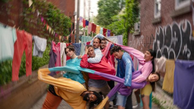 in an alley lush with vegetation, eight dancers are tangled together both with their limbs and with the sleeves of their brightly colored clothing; a slight motion blur on the edges of the frame suggest the camera itself is spinning