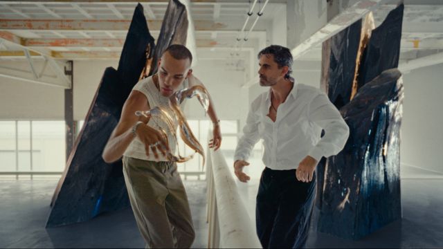 two men posture in a room with large crystal like structures growing unnaturally out of the floor; one man seems to be conjuring some metallic fluid with his jewelry-clad right hand