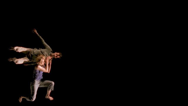 on a solid black background, a light skinned dancer in a purple shirt is down on one knee and holding another dancer effortlessly on her left shoulder