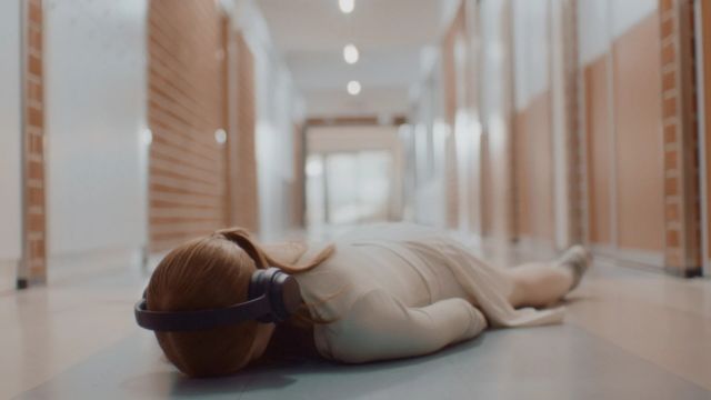 a girl with long red hair lies face down on the floor of an otherwise empty school hallway; she is wearing headphones