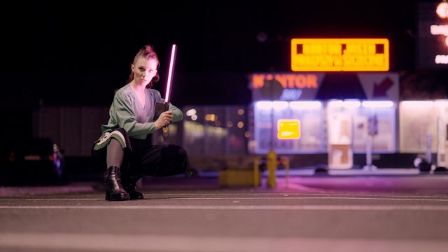 a young, fair-skinned woman squats on pavement at night, holding a neon pink strip of LEDs in what appears to be a parking lot with illuminated signs behind her