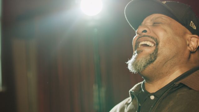 a closeup of a light brown skinned man wearing a hat, laughing boisterously with a stage light behind him