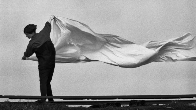 a black and white image of a dancer with short dark hair holding a long sheet of fabric - several times the length of their body - off the ground as the wind ripples through it
