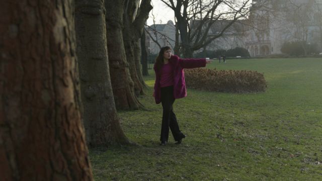 a young woman in black pants and a magenta fur coat strolls along a row of large trees in front of an impressive multi-story stone building