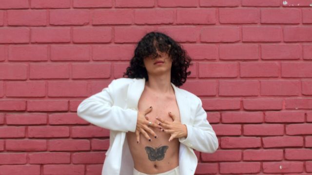 in front of a brick wall painted red, a dancer with dark curly hair covering their eyes and a butterfly tattoo above their bellybutton poses, white jacket open exposing a bare chest