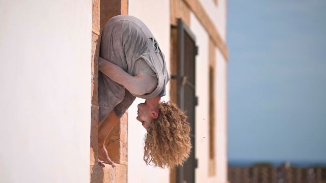 a dancer stands in a windowsill, leaning forward and out of the window, their wavy hair hanging down