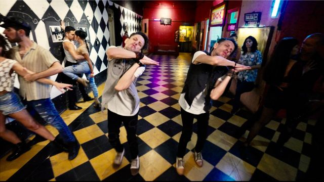 in a theater lobby painted in a checkerboard pattern, two twins pose identically with palms touching as several couples dance as partners behind them