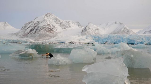 in a shallow inlet filled with chunks of sea ice, a woman in a black dress stands in waist deep water, bending forward with left arm extended as her hair touches the surface