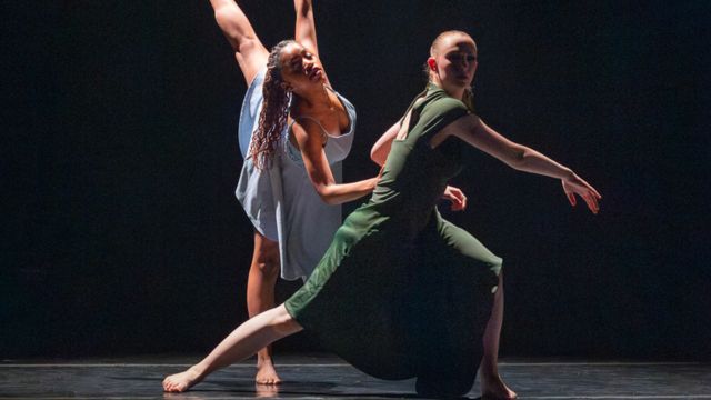in an all black room, a dancer with fair skin in a long green dress extends her right leg backward; behind her, a dark skinned dancer with long brown hair extends her left leg toward the ceiling