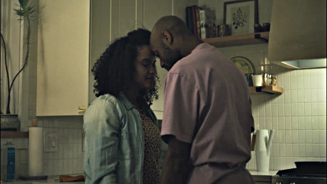 a dark skinned husband and wife touch their foreheads together while standing in a residential kitchen with white cupboards and tile walls