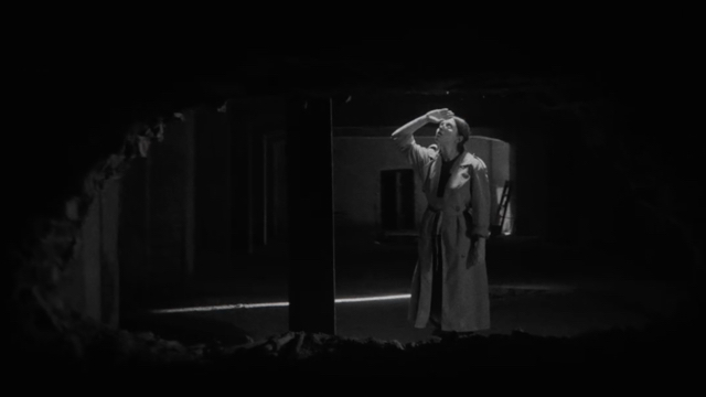 through a hole in the wall, we see a light skinned woman in a trench coat looking upward, hand shielding her eyes from a bright light source