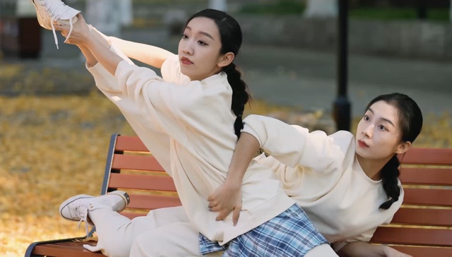 two Asian women pose on a park bench; one is holding the other's right ankle high in the air