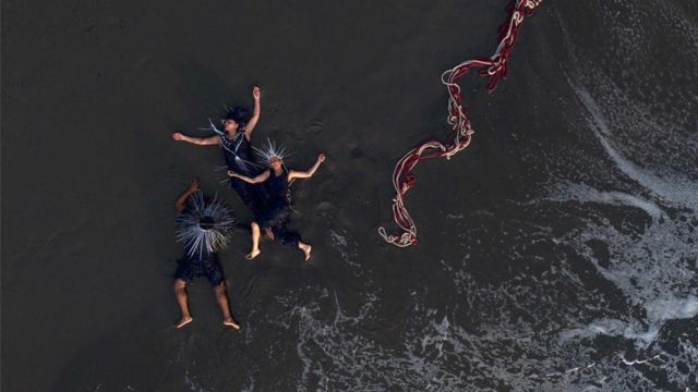 three dancers wearing spiked urchin-like crowns lay on a wet beach alongside a tangle of red and white rope