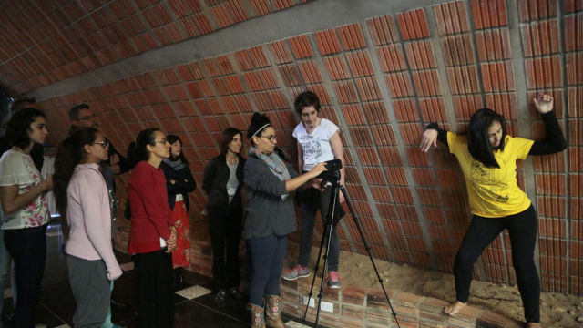 a dancer presses herself against a wall while a workshop facilitator and several participants watch from behind a camera on a tripod