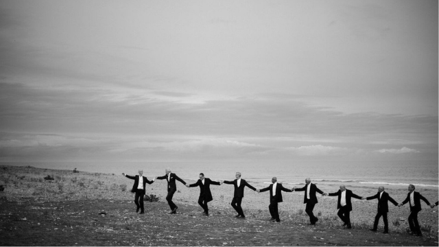 a monochrome shot of ten men in black suits holding hands and walking along a barren landscape