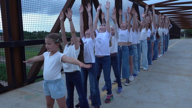 a large group of children in jeans and white tee shirts stand in a line on a bridge, arms extended above their heads