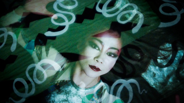 a woman looks with intense eyes at the camera; her face is partially obscured by looping patterns drawn on the glass in front of her