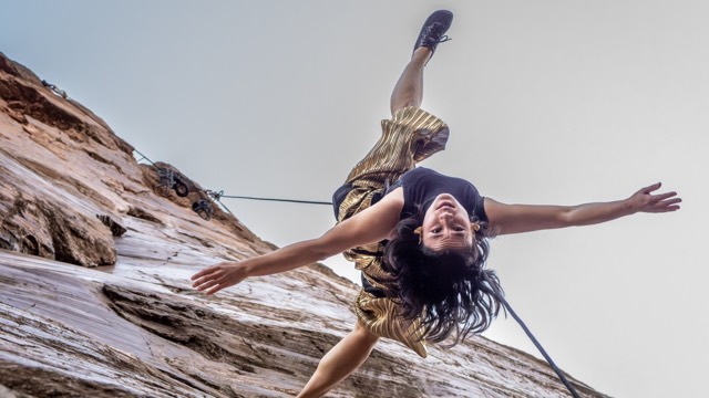 a dark haired dancer - suspended upside down from a cable on a sheer rock wall - looks at the camera, smiling